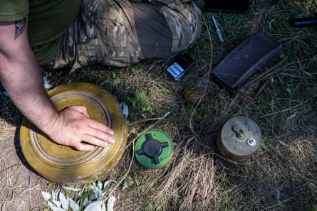 The hand of a man wearing combat trousers rests on a shallow cylindrical object the size of a dinner plate. Other devices can also be seen on the ground.