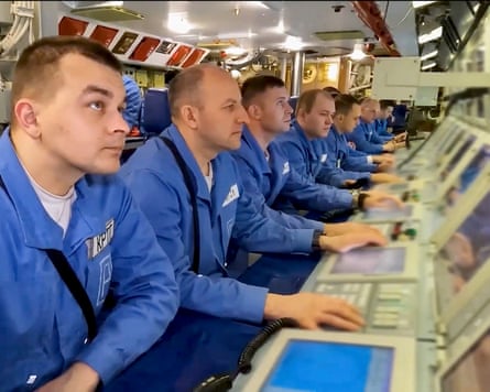 Submarine crew dressed in blue uniforms sit at a bank of monitors