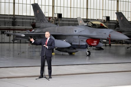 A man stands in front of a fighter jet in an aircraft hangar.