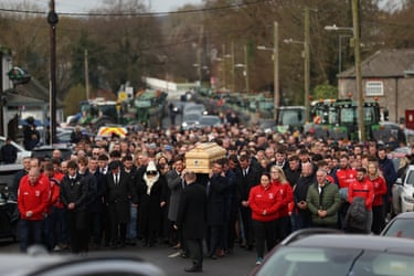 A large group of mourners surround a coffin carried through the street 