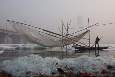 A fisher stands on a wooden boat next to a big fishing net covered with foam in the polluted Yamuna river on a smoggy morning