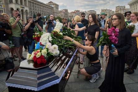 People lay flowers on a coffin in a city square.