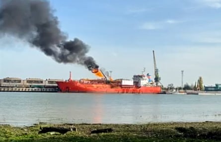 A pillar of flames and black smoke rises at an angle into the blue sky from the red-hulled cargo ship moored at the opposite side of the river