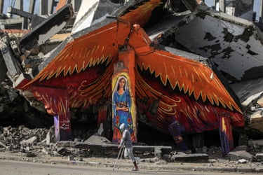 A man walking with crutches passes a mural of a woman in a blue robe under what appear to be the wings of a bird