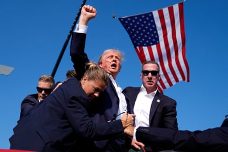 Republican presidential candidate former President Donald Trump, with blood on his face, is surrounded by US Secret Service agents, an American flag flying behind them. after an assassination attempt at a campaign rally in Butler, Pennsylvania, July 13 2024