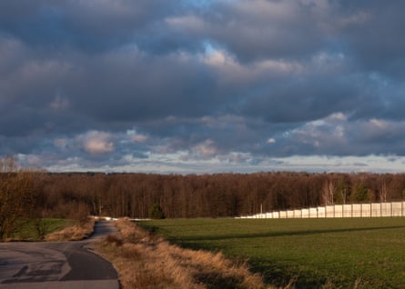 Landscape of sloping fields with forest in the background under a cloudy sky; a border fence can be seen in the background. This is the border fence in the Podlasie region