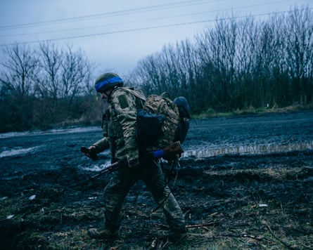 A soldier is wearing a helmet with blue tape wrapped around it. He has a large backpack on his back and is holding a gun