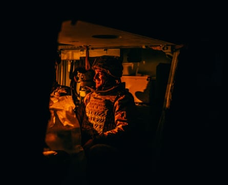 A soldier sitting in the armoured vehicle is smiling, lit by the glow of the light in the vehicle