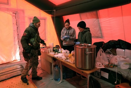A man in camouflage gear with two women behind a table with a giant cooking pot.