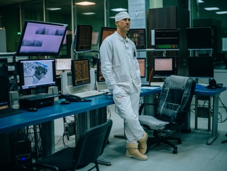 A worker in white overalls and a white hat during his shift at Chornobyl. Screens showing security camera footage sit on a desk behind him