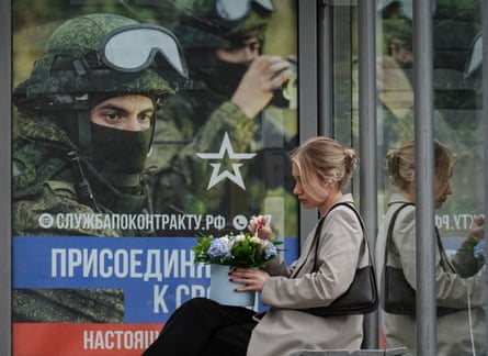 A woman sits at a bus stop in Moscow, in front of a poster advertising military conscription.