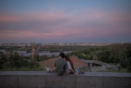 A man and a woman sit together on a wall, she with her legs over his lap, before a cityscape with pink clouds