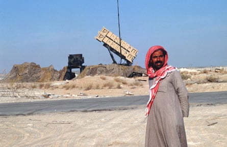 A Saudi ma nin red headscarf walks in the foreground with a Patriot missile defence system behind