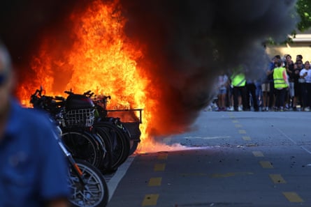 Dense flames rise from a vehicle on fire as police hold back a group of protesters in the background. 