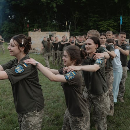 Cadets dance and sing around a bonfire: they are in a line with their hands on one another’s shoulders as if in a conga. They are casually dressed in short-sleeved khaki shirts with badges on the sleeves and camouflage combat trousers. The girls have their hair tied back.