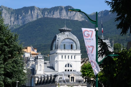 A G7 flag flies in front of a large white building with a tall tower in Evian-les-Bains, France. There are mountains in the background.