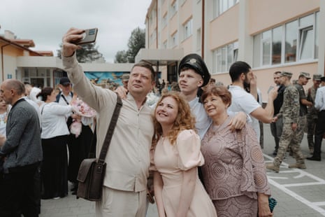 A family takes a selfie with a graduating cadet: a young man in a black cap and white shirt stands with his arm around an older lady in a long-sleeved light brown lace top and skirt and behind a woman with reddish-blond curly hair who may be his mother, who wears a peach-coloured dress with puffy short sleeves, while a man who may be his father, wearing a pale cream shirt and trousers, holds up a camera.