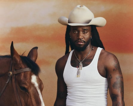 A Black man in a big cream coloured cowboy hat and a white vest stands next to a horse. 