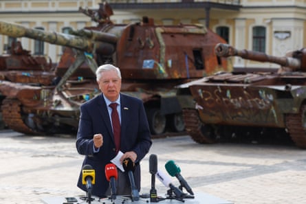 lindsey graham, an older man in suit, speaks into microphones outside with damaged tanks behind him 