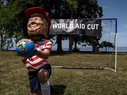 a person wearing large head mark of Donald Trump kneels in front of a soccer goal that says ‘world aid cut’