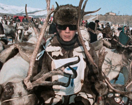 A man wearing a fur hat and dark sunglasses carrying a dead moose