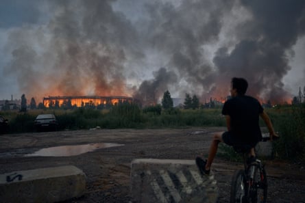 A teenager sits on a bicycle watching flames rise from a burning logistics warehouse.