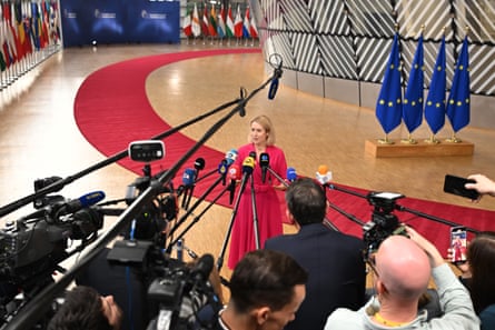 Kaja Kallas in red dress surrounded by reporters with boom mics hanging overhead.