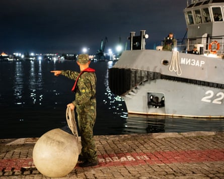 A Bulgarian navy sailor points while holding rope, with the Mizar rescue tug moored nearby at night