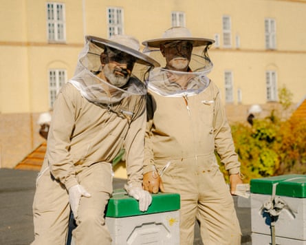 Two beekeepers in beige protective clothing outdoors, with a yellow building behind them.